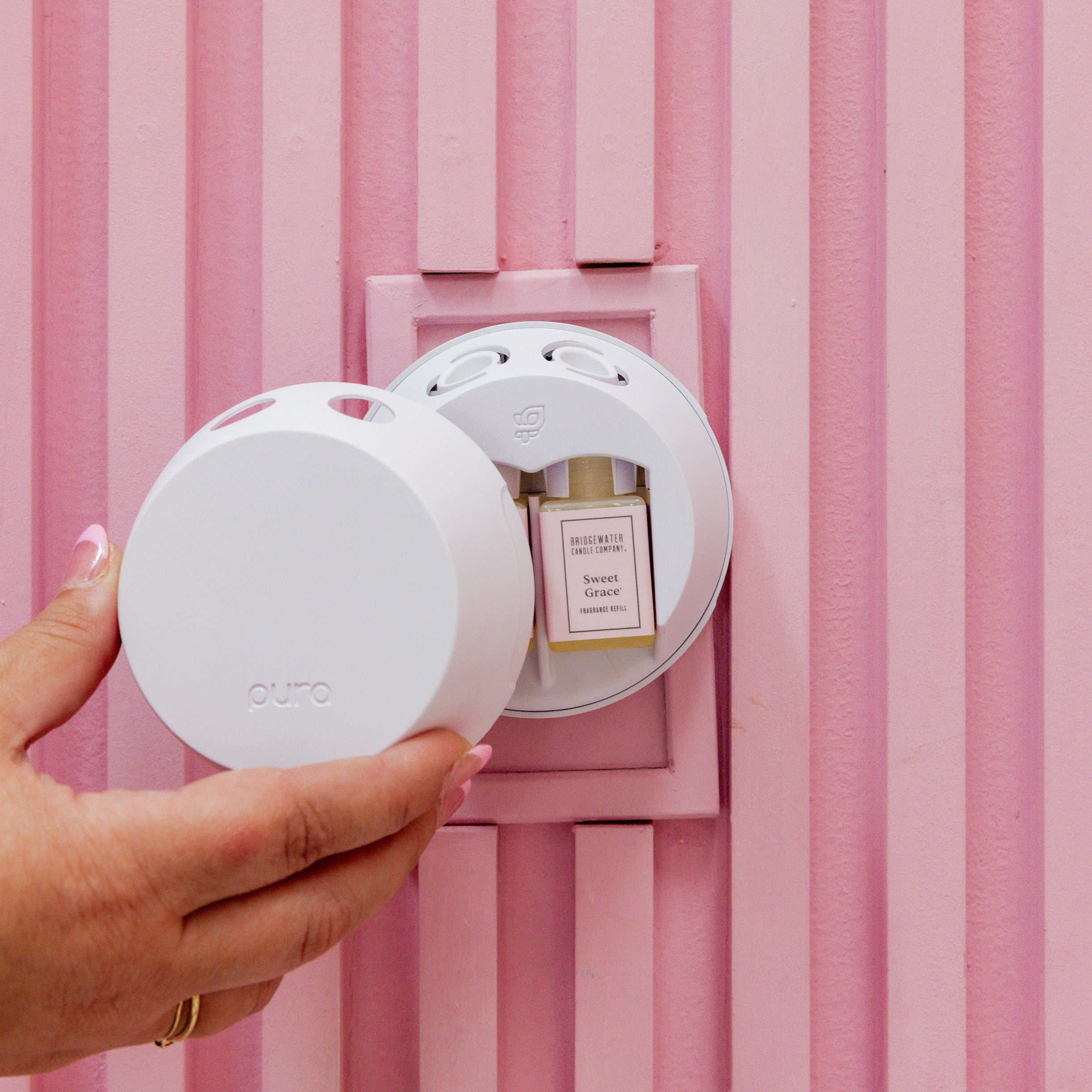 A hand inserts the white Bridgewater Candle PURA + Bridgewater Smart Home Diffuser into a pink wall outlet, beside a Sweet Grace fragrance vial. The background displays vertical pink paneling.
