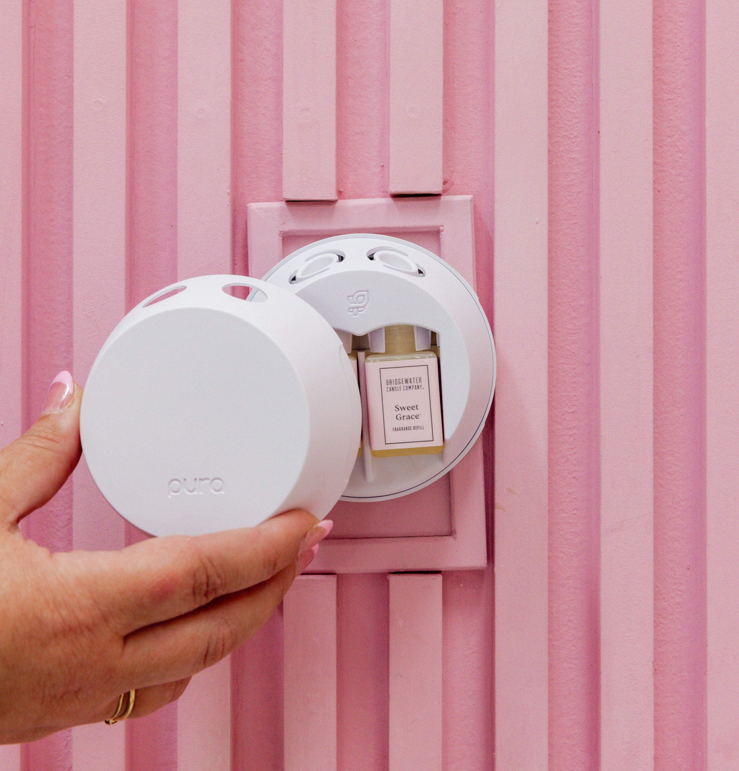 A hand inserts the white Bridgewater Candle PURA + Bridgewater Smart Home Diffuser into a pink wall outlet, beside a Sweet Grace fragrance vial. The background displays vertical pink paneling.