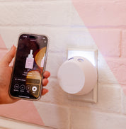 A hand holds a smartphone displaying an app next to a plugged-in PURA + Bridgewater Smart Home Diffuser Set with Sweet Grace and Afternoon Retreat V5 from Bridgewater Candle, glowing softly while mounted on a white wall with pink geometric designs.