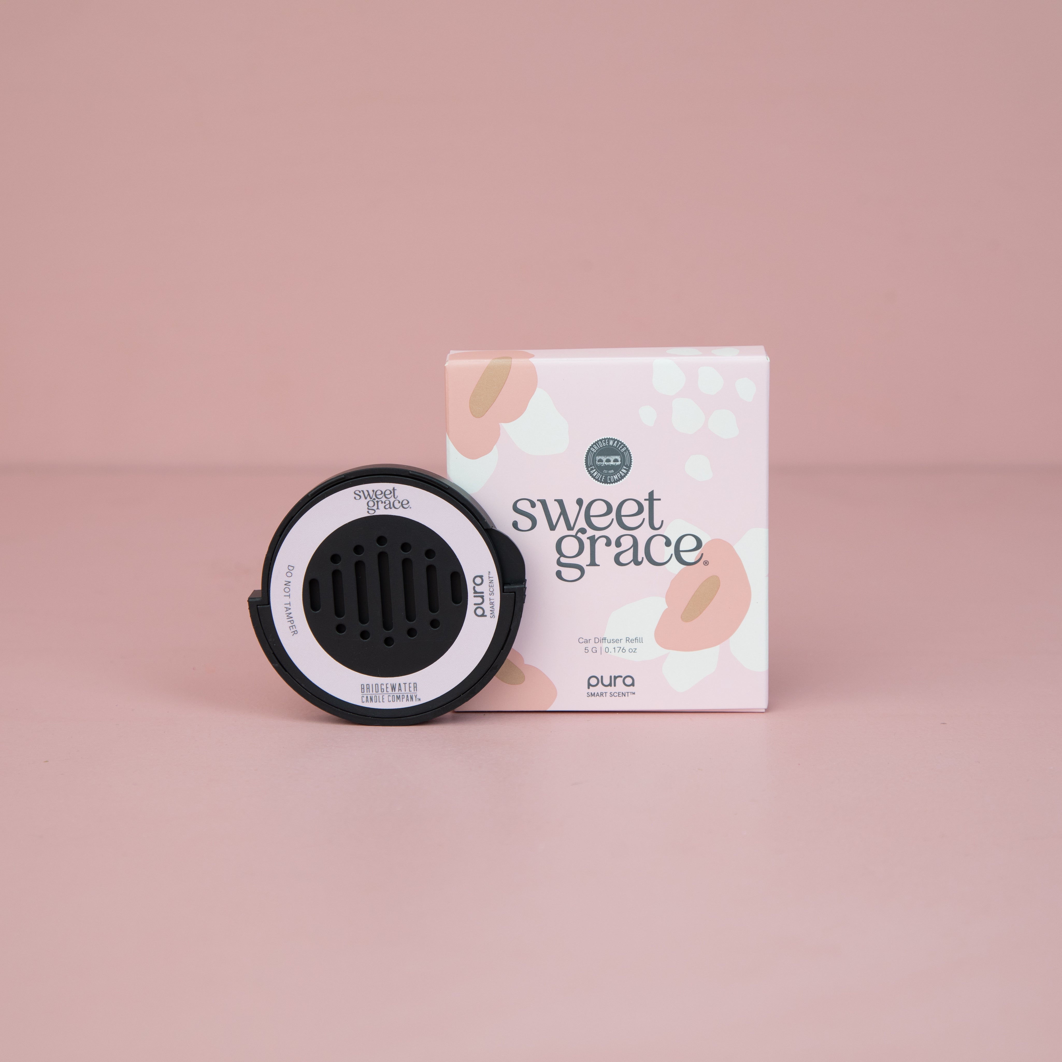 A Bridgewater + Pura Car Diffuser Cartridge Sweet Grace by Bridgewater Candle Company sits before a pastel pink and white box labeled sweet grace and pura, against a light pink background—ideal as a car air freshener.
