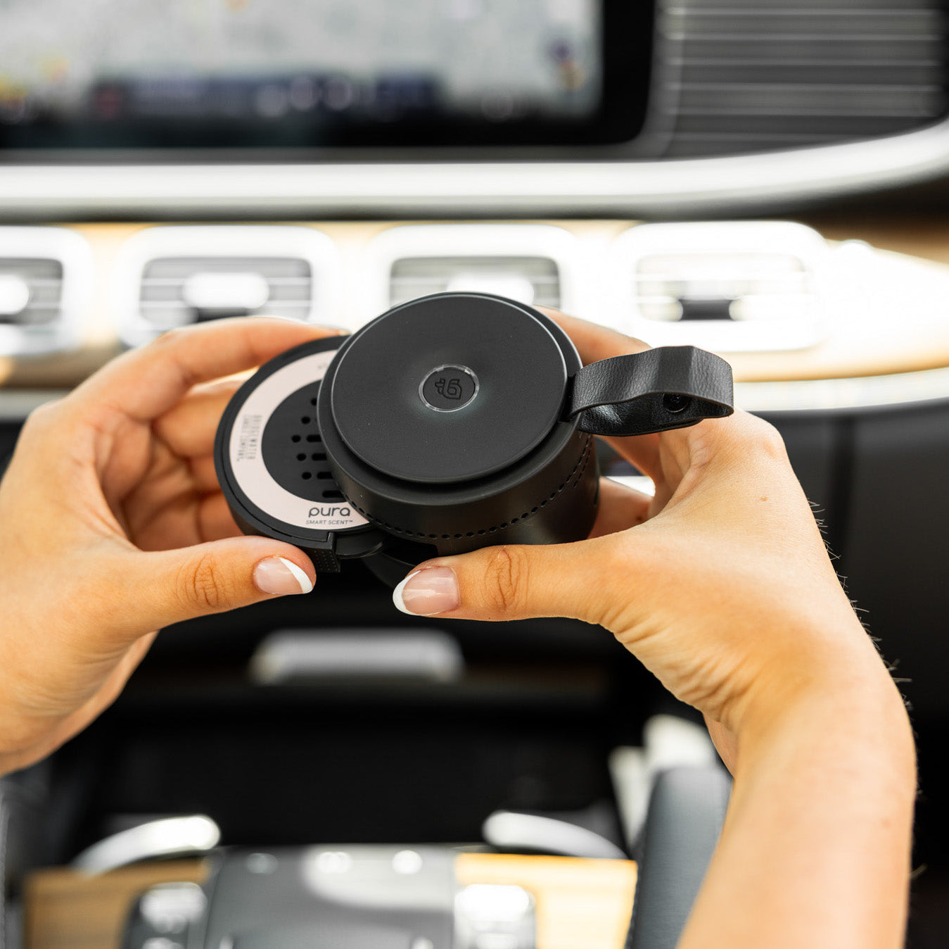 Someone holds the round, black Bridgewater + PURA Sweet Grace Car Diffuser Set by Bridgewater Candle Company inside a car, with the dashboard and controls visible in the background.