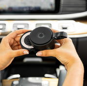 Someone holds the round, black Bridgewater + PURA Sweet Grace Car Diffuser Set by Bridgewater Candle Company inside a car, with the dashboard and controls visible in the background.