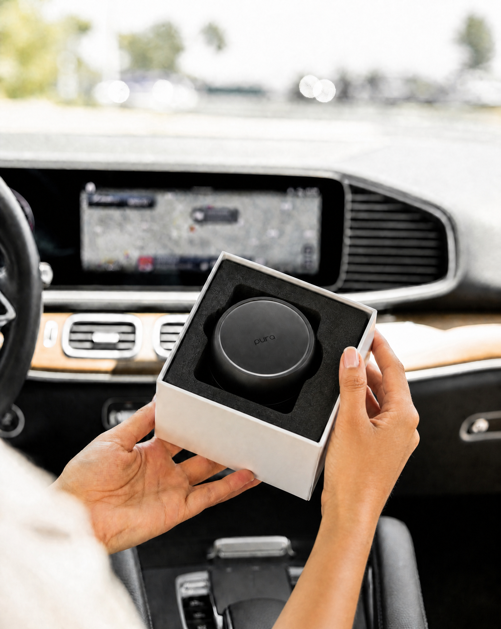 A person holds an open box with a round, black Bridgewater + PURA Sweet Grace Car PRO Diffuser Set by Bridgewater Candle inside a car; “pura” is visible on the diffuser, and a navigation screen is seen in the background.