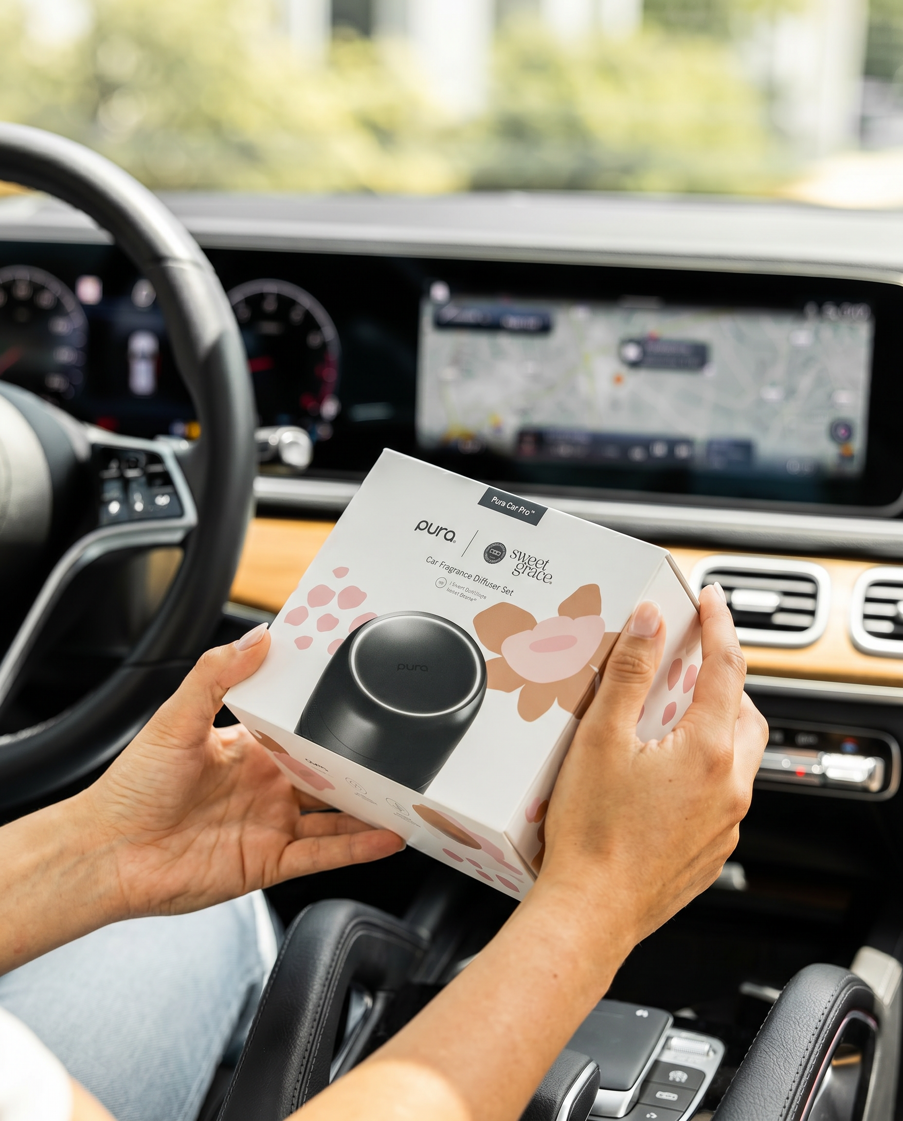 A person in a car holds the Bridgewater + PURA Sweet Grace Car PRO Diffuser Set by Bridgewater Candle, with the dashboard, steering wheel, and screen in view—ready to enjoy lasting fragrance from this smart car diffuser.