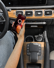 A person in a car holds a smartphone showing a charging screen above the wood center console, next to a Bridgewater + PURA Sweet Grace Car PRO Diffuser Set by Bridgewater Candle for long-lasting fragrance.