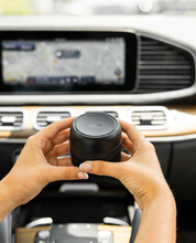 A person holds the Bridgewater + PURA Sweet Grace Car PRO Diffuser Set by Bridgewater Candle inside a car, with a GPS screen in the background, suggesting convenient long-lasting fragrance while driving.