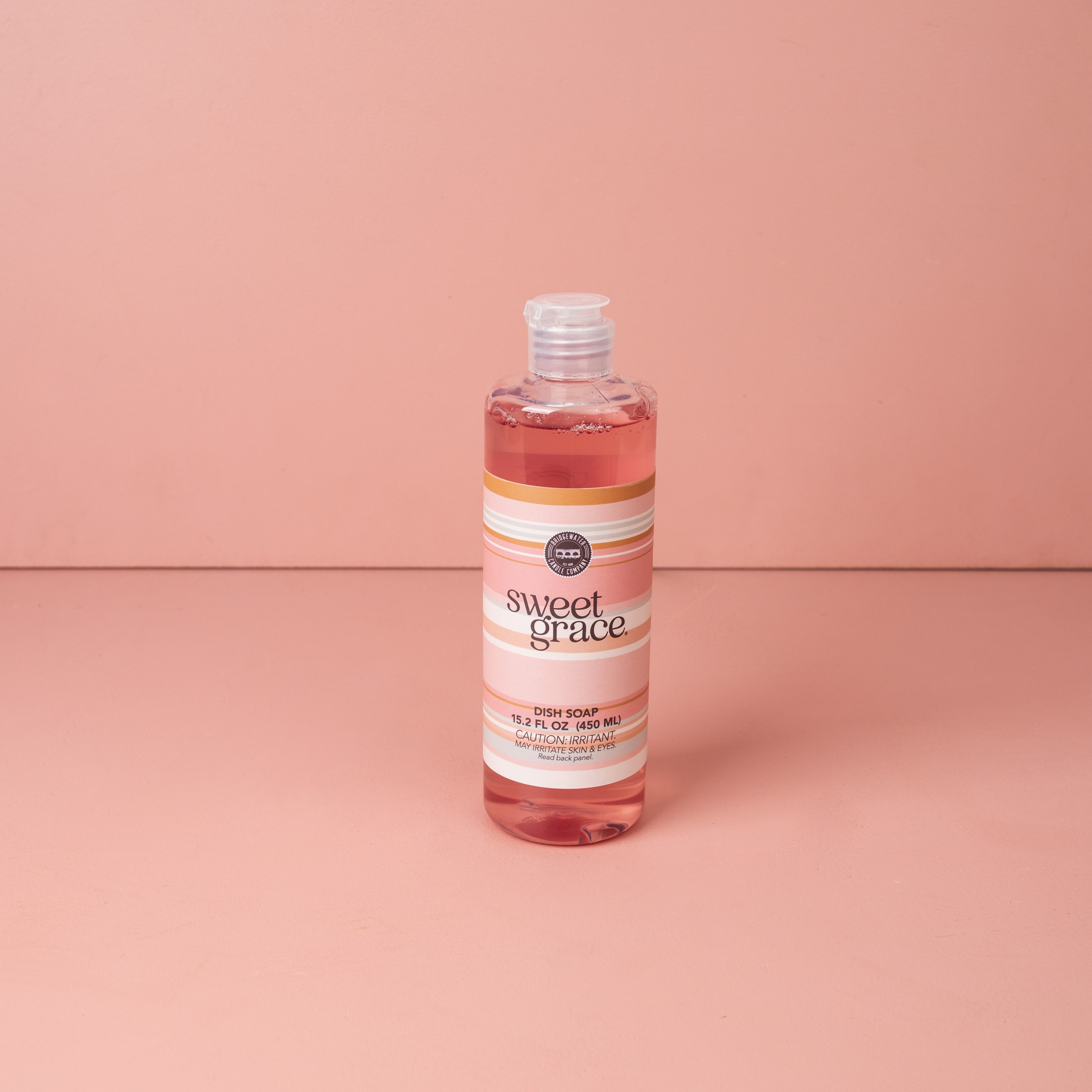 A clear bottle of Bridgewater Candle Company’s Sweet Grace Dish Soap, with a white label and pink accents, stands on a pink surface against a pink background. The cap is open, showcasing its best-selling fragrance.