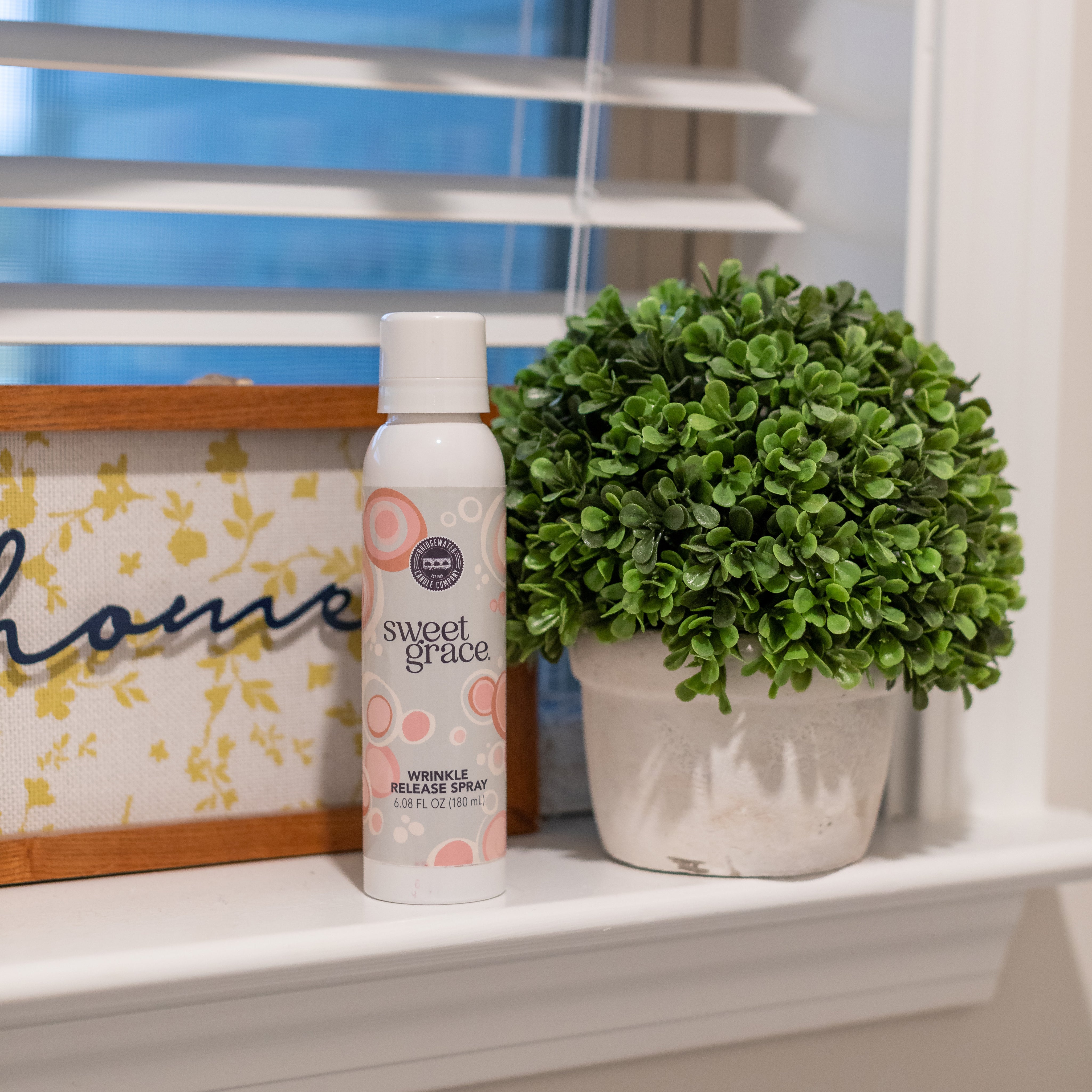 A can of Sweet Grace wrinkle release spray sits on a windowsill next to a potted artificial plant and a decorative home sign with white blinds in the background.