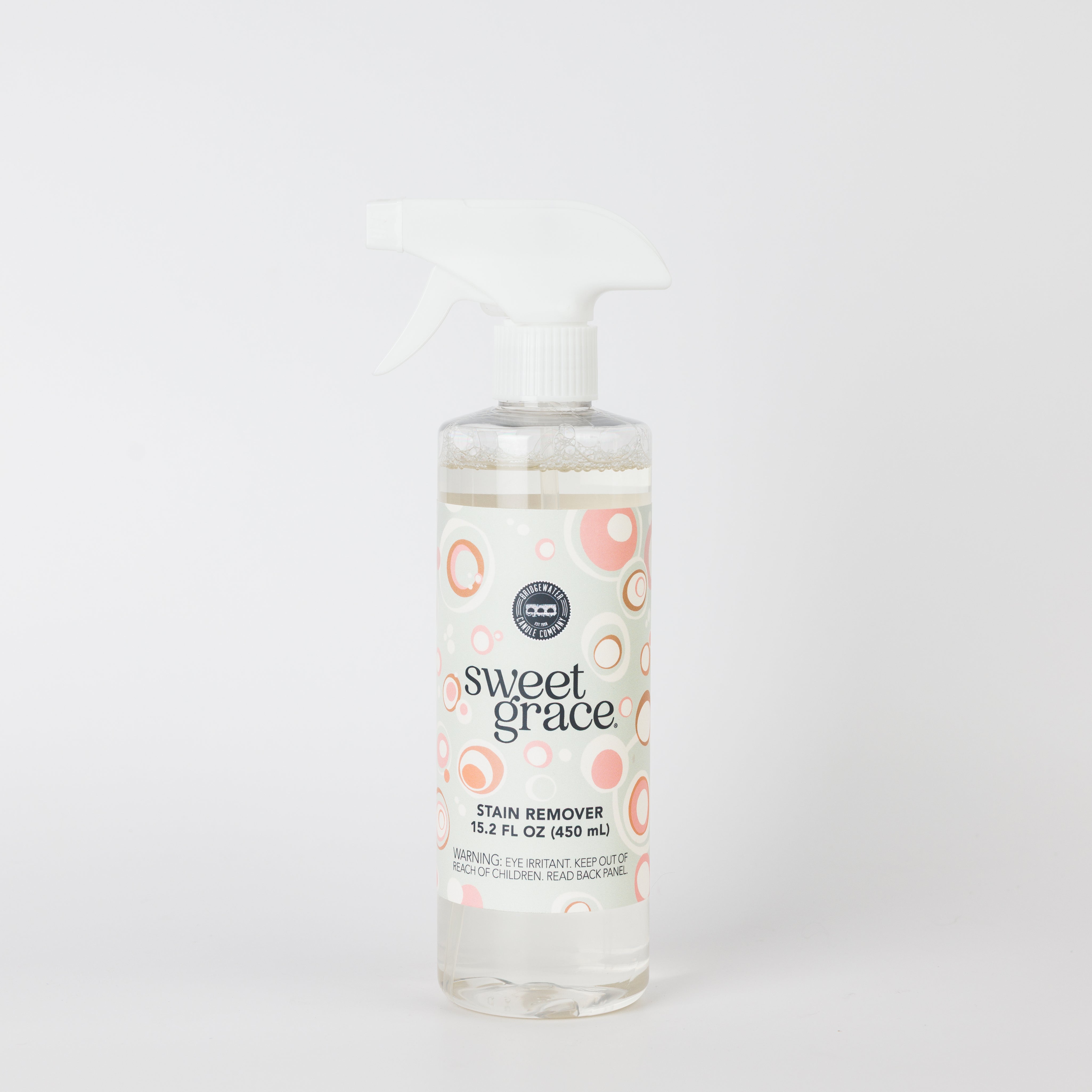 A clear plastic spray bottle of Bridgewater Candle’s Sweet Grace Stain Remover with a white trigger and a pink, orange, and beige circle-patterned label stands against a white background. Safe for most fabrics.