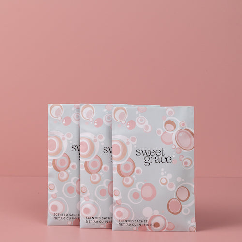 Three Sweet Grace scented sachets with white and pink circular patterns stand upright against a solid pink background.