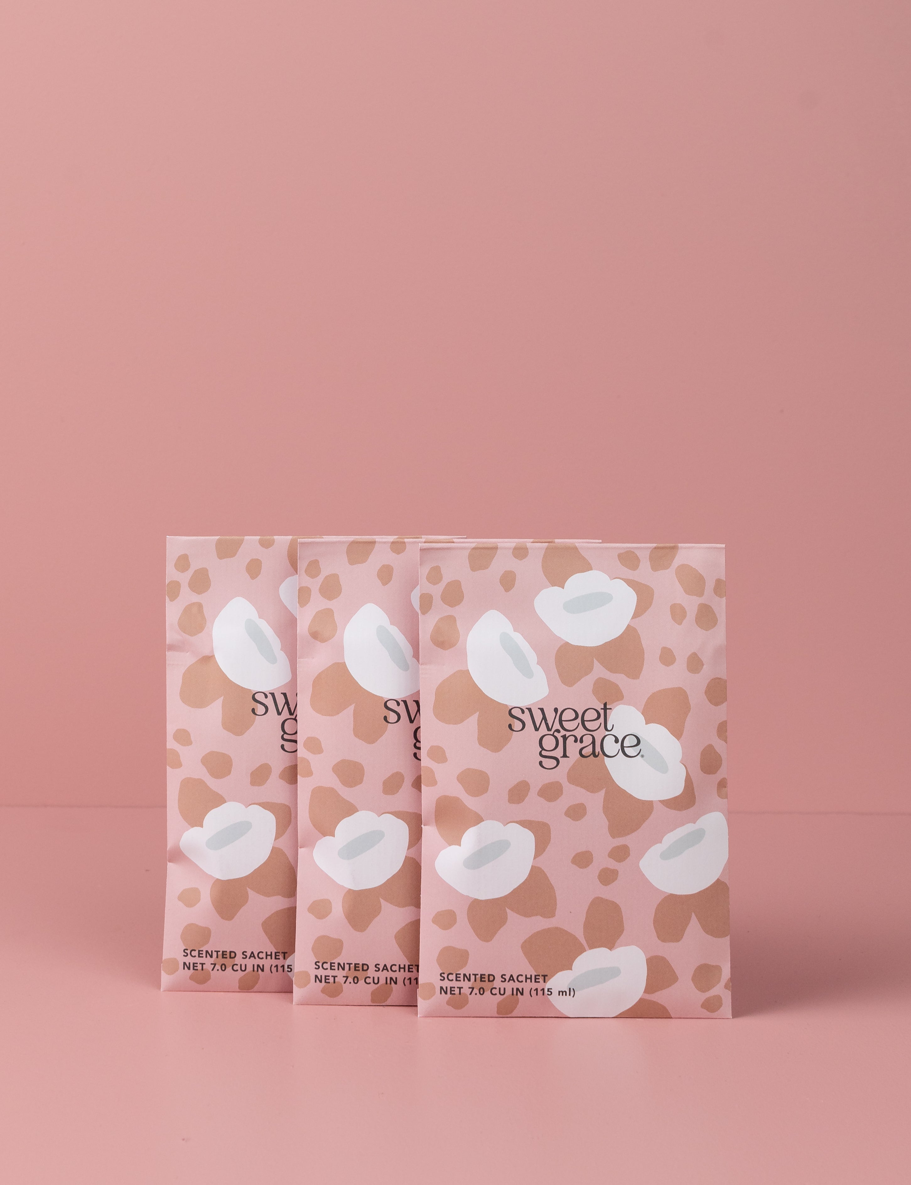 Three Sweet Grace Modern Flower Sachets by Bridgewater Candle Company, featuring pink and beige floral patterns, stand upright against a matching background—adding the beloved Sweet Grace fragrance to your home collection.
