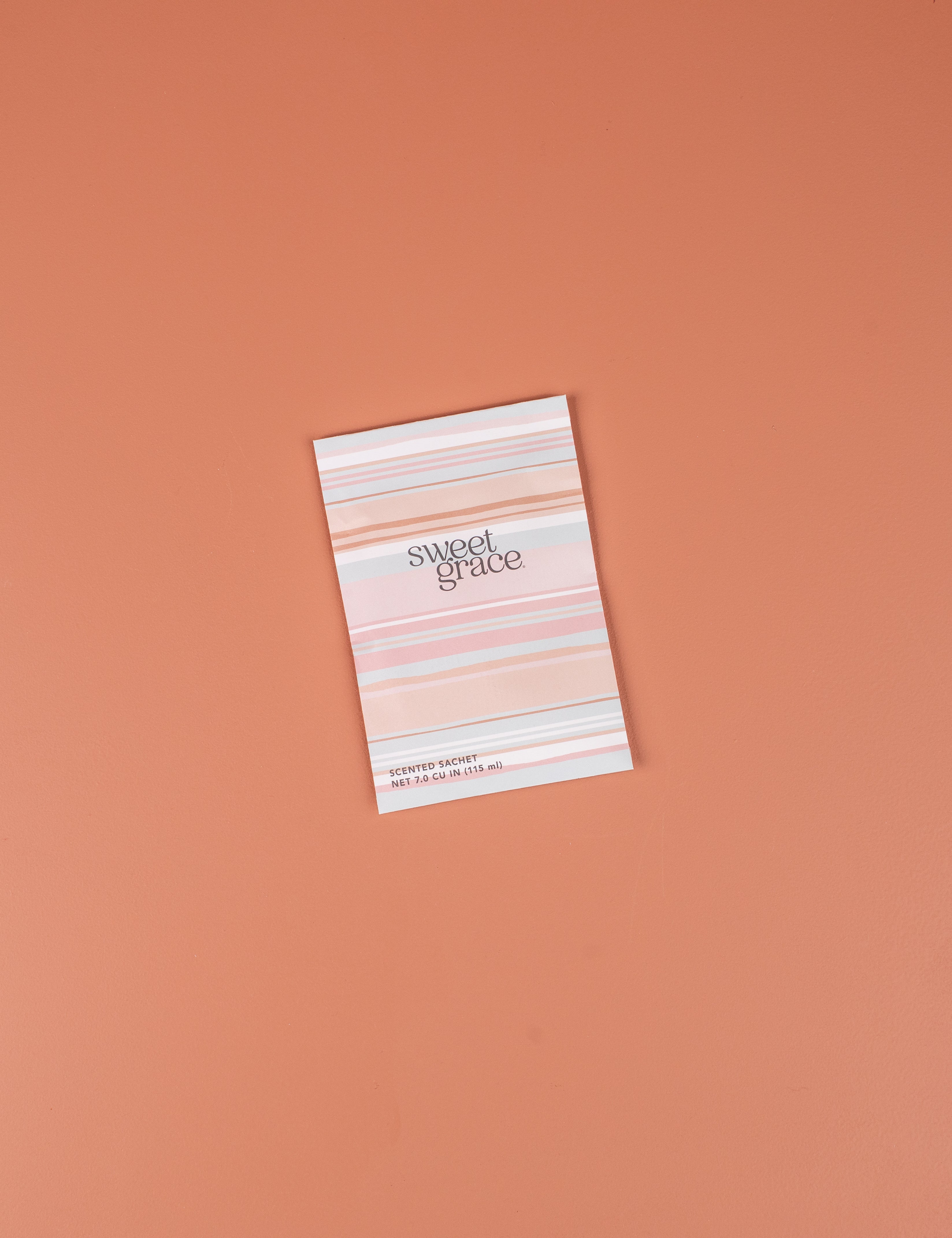 The Bridgewater Candle Company Sweet Grace Modern Stripe Sachet, featuring a pink and white striped design, rests on a peach surface—ideal for infusing your space with home fragrance.