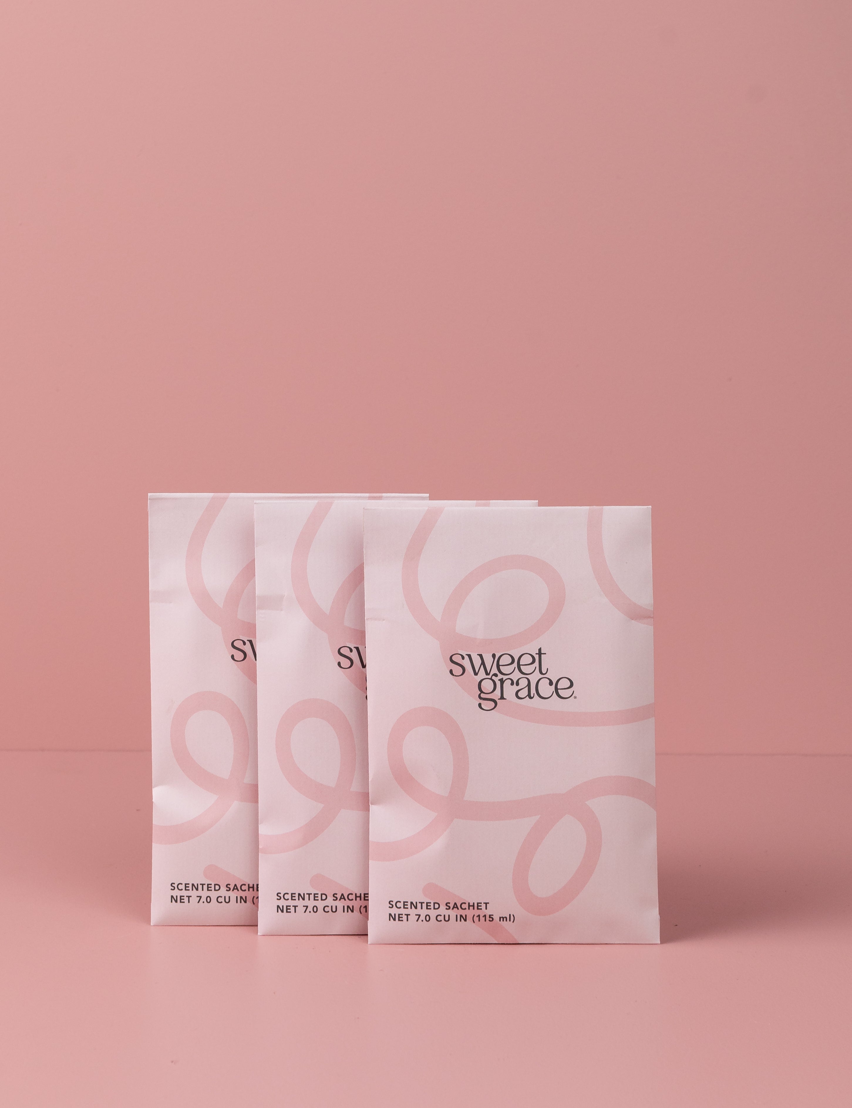 Three Sweet Grace Modern Swirl Sachets by Bridgewater Candle Company, in pastel pink with elegant swirls and typography, are arranged side by side on a matching background—perfect for adding a fragrant touch to any space.