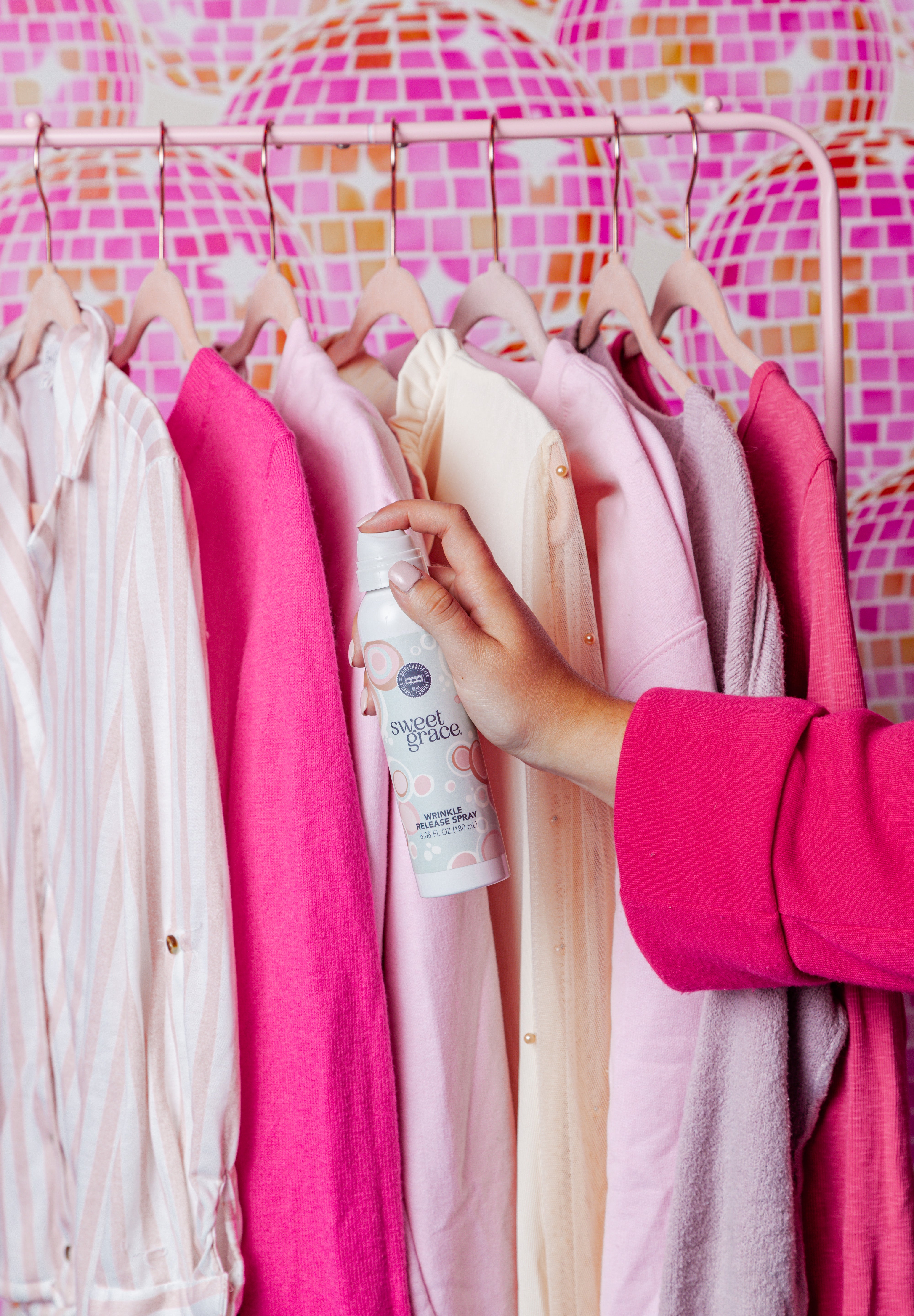 A person in a bright pink sleeve holds a can of Sweet Scent deodorant spray near a rack of pink and cream shirts against a colorful pink mosaic background.