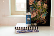 A silver bottle of Bridgewater Candle Company’s Nantucket Coast Non-Aerosol Room Spray rests on two stacked books, with a vibrant floral painting behind them on a white countertop near a window.