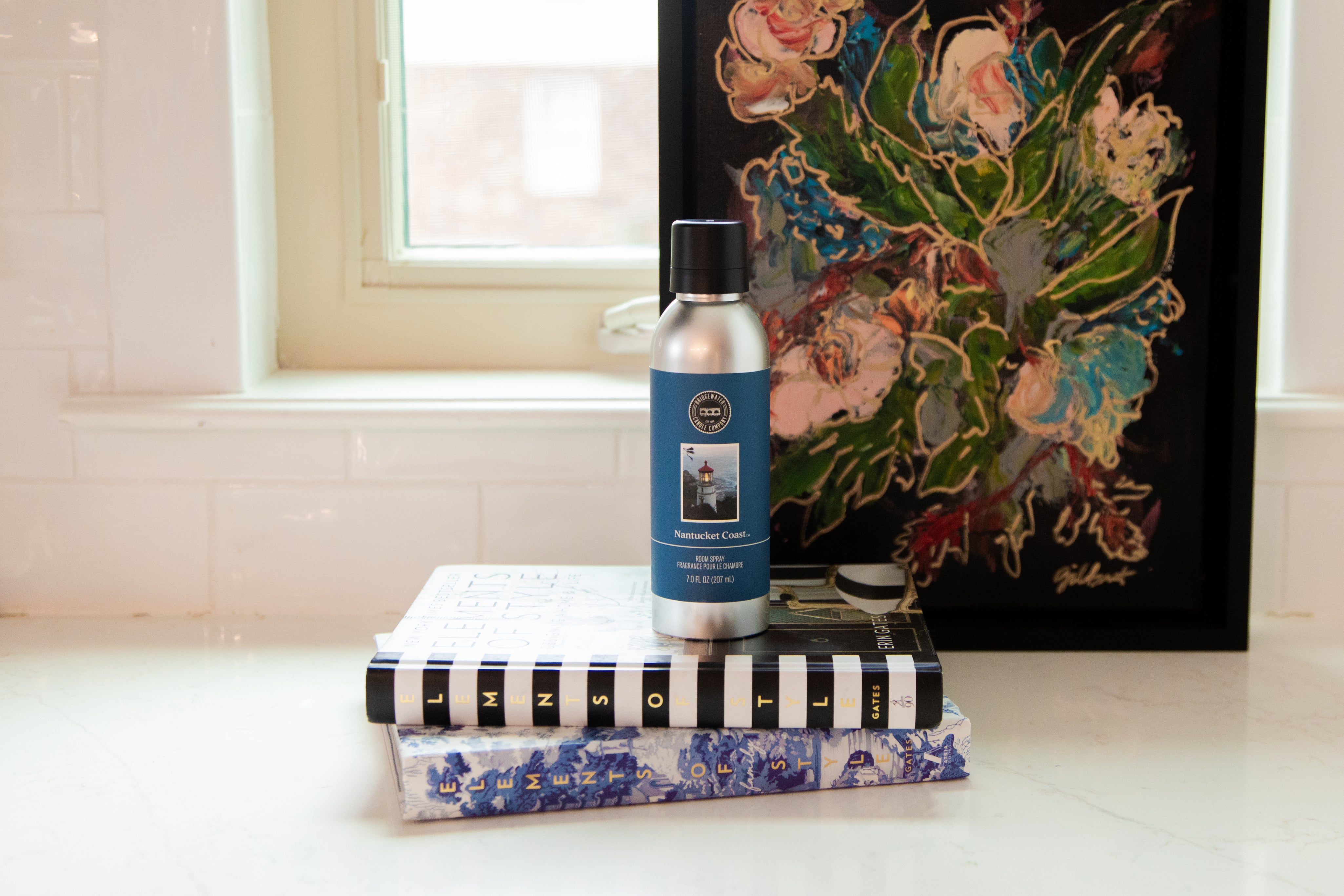A silver bottle of Bridgewater Candle Company’s Nantucket Coast Non-Aerosol Room Spray rests on two stacked books, with a vibrant floral painting behind them on a white countertop near a window.