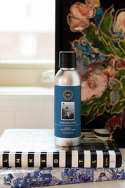 A silver Nantucket Coast Non-Aerosol Room Spray by Bridgewater Candle Company rests on books by a window and floral art, featuring a lighthouse image—an eco-friendly way to refresh your homes scent.