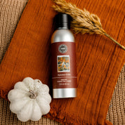 A bottle of Bridgewater Candle’s Autumn Gold Room Spray sits on a rust-colored cloth next to a white pumpkin and dried wheat stalks, creating a cozy fall-inspired home fragrance display.