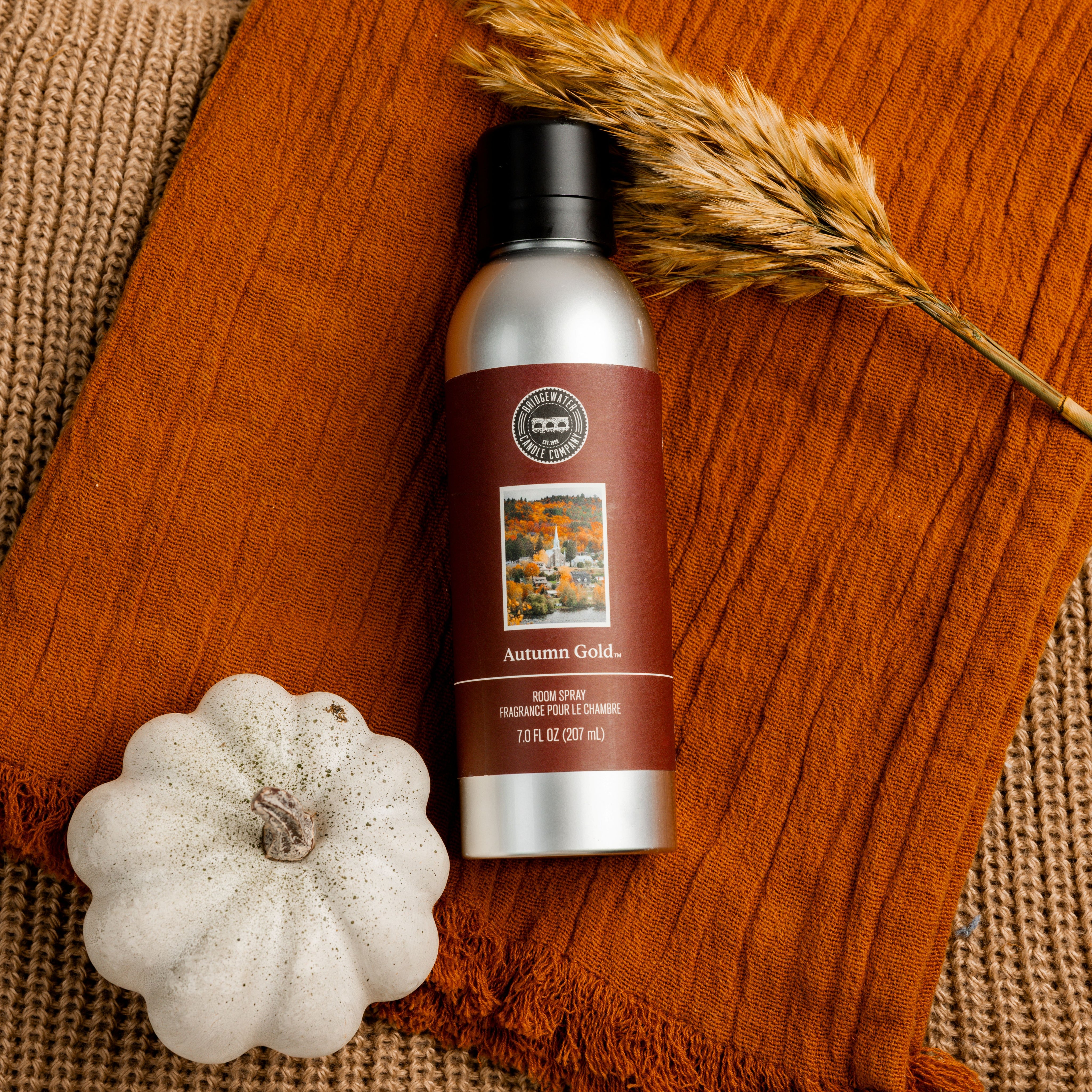A bottle of Bridgewater Candle’s Autumn Gold Room Spray sits on a rust-colored cloth next to a white pumpkin and dried wheat stalks, creating a cozy fall-inspired home fragrance display.