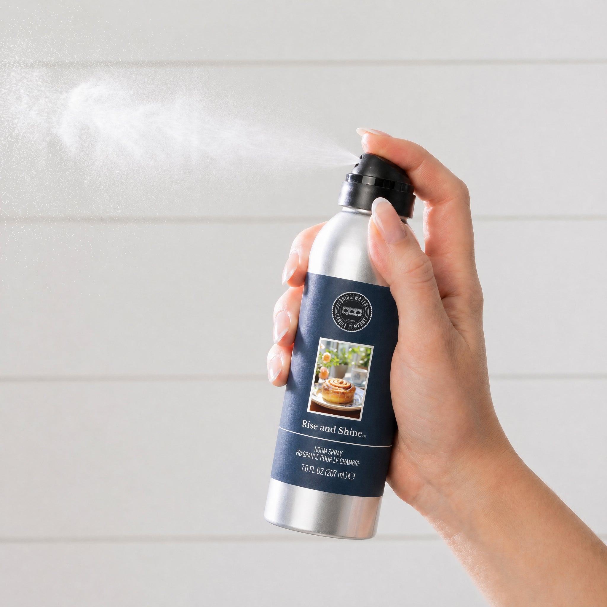 A hand sprays Bridgewater Candle’s Rise & Shine Non-Aerosol Room Spray, shown in a silver bottle with pancake artwork. The light, softly blurred background highlights this eco-friendly room fragrance.