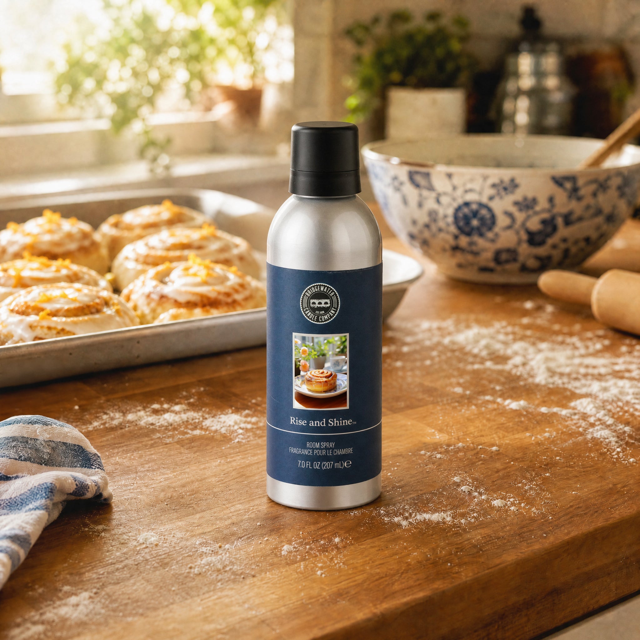 A can of Bridgewater Candle Rise & Shine Non-Aerosol Room Spray sits on a floured kitchen counter with pastries, a patterned bowl, and sunlight—filling the air with its cozy cinnamon roll fragrance.