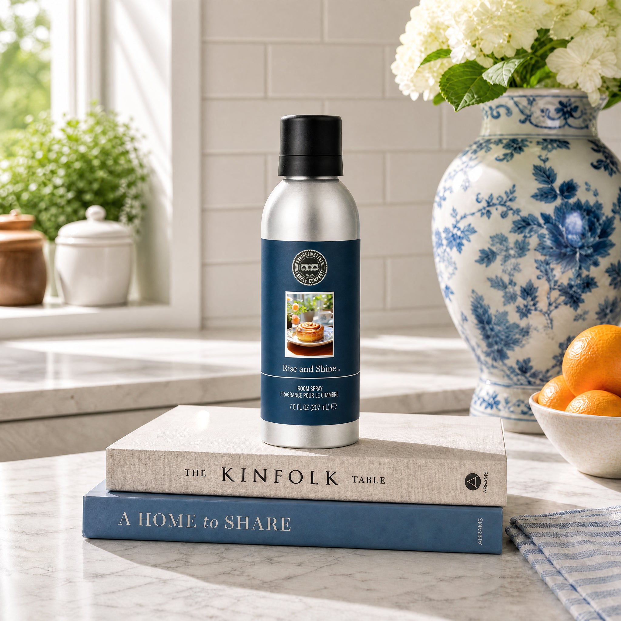 A silver Rise & Shine Non-Aerosol Room Spray by Bridgewater Candle sits on two stacked books atop a marble counter, beside a blue-and-white floral vase and a bowl of oranges in a sunlit kitchen—an eco-friendly room fragrance.