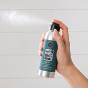 A hand mists Bridgewater Candle’s “I’ll Be Home” Non-Aerosol Room Spray, featuring a silver bottle with a dark green label. The soft, light background enhances this cozy, eco-friendly home fragrance’s holiday appeal.