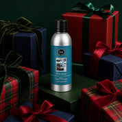 The Bridgewater Candle "I'll Be Home" Non-Aerosol Room Spray, in a sleek silver bottle, is displayed among festively wrapped red, green, and plaid gifts with ribbon bows against a dark green backdrop—a cheerful accent for any room.