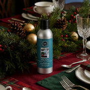A silver bottle of Bridgewater Candle’s I’ll Be Home Non-Aerosol Room Spray sits on a holiday table decorated with pine branches, pinecones, gold ornaments, and red plaid, surrounded by neatly arranged plates and glassware.