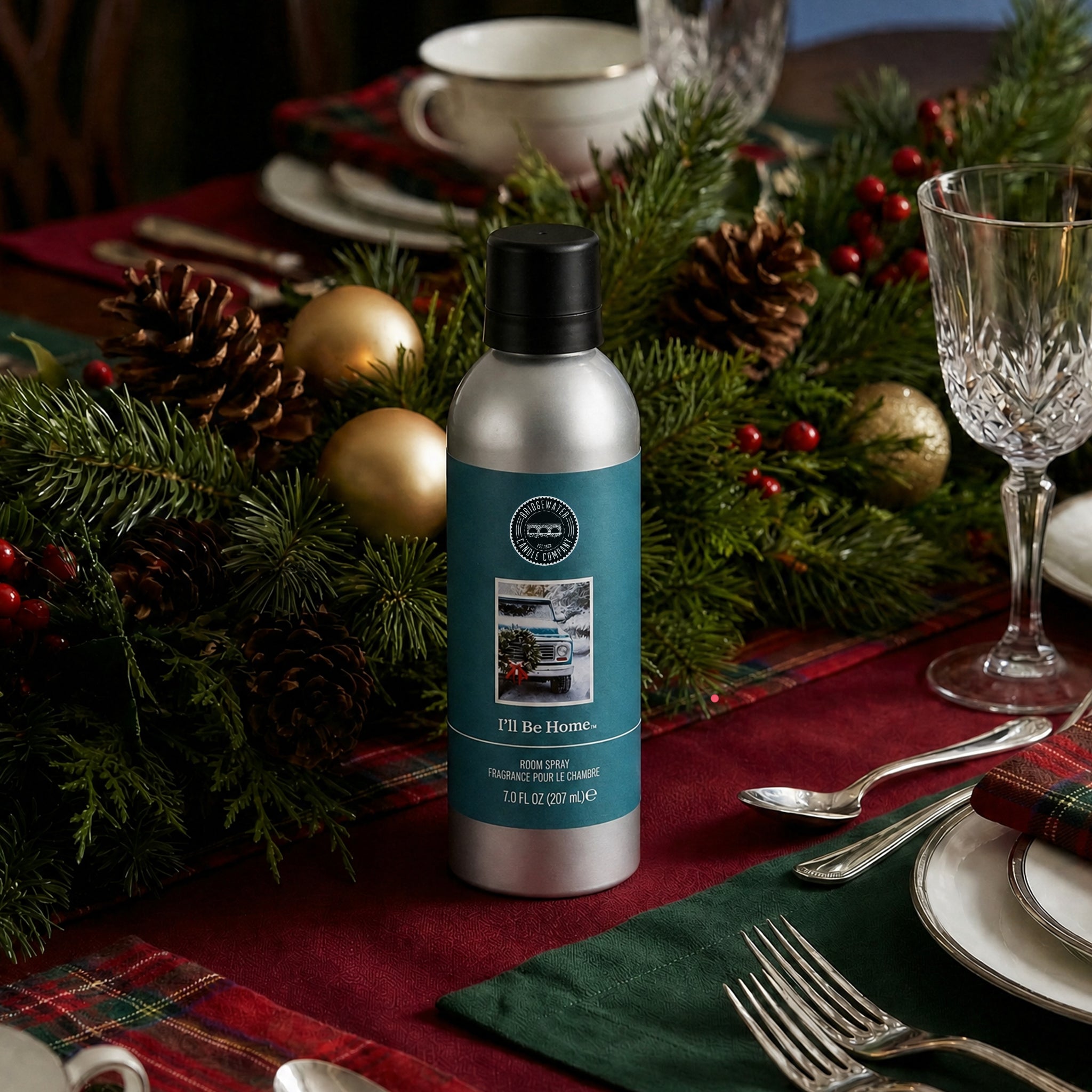 A silver bottle of Bridgewater Candle’s I’ll Be Home Non-Aerosol Room Spray sits on a holiday table decorated with pine branches, pinecones, gold ornaments, and red plaid, surrounded by neatly arranged plates and glassware.