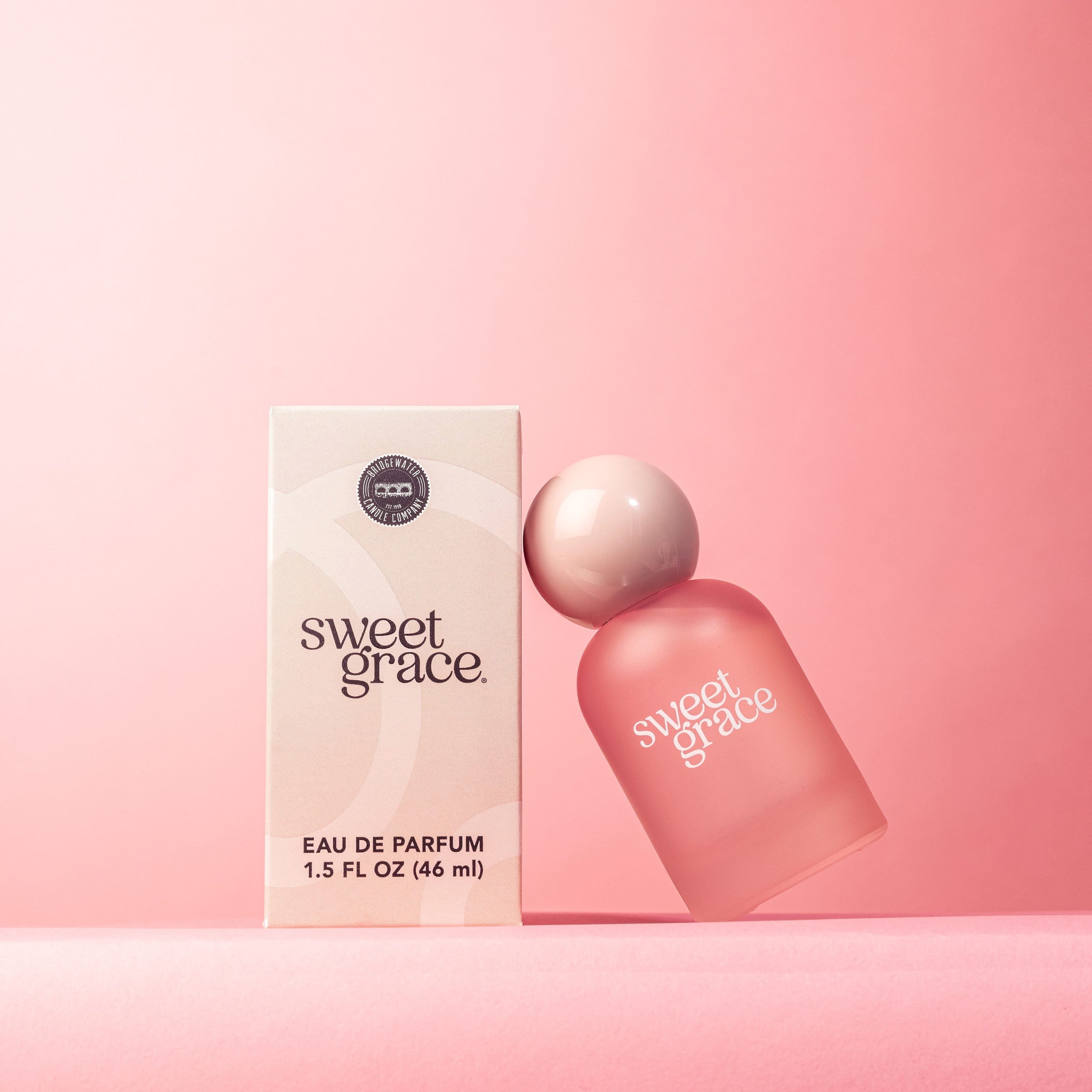 A frosted pink bottle of Sweet Grace Eau de Parfum with a glossy pink ball top, displayed next to its packaging box on a pink background.