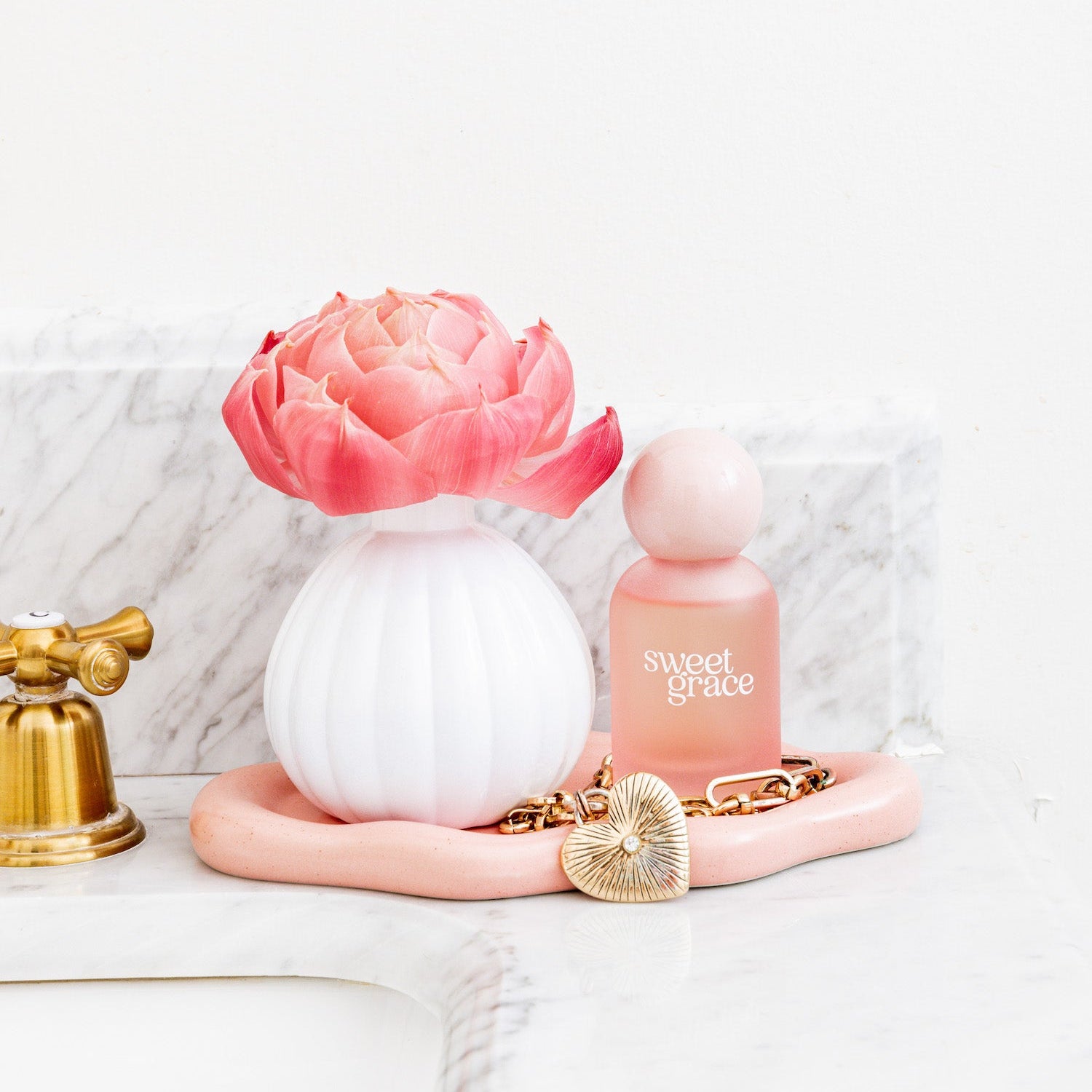 The Bridgewater Candle Company Sweet Grace Flower Diffuser sits on a pink tray with a gold chain and heart locket, placed on a marble bathroom counter near a gold faucet.