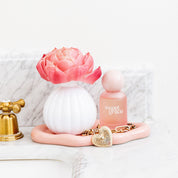 The Bridgewater Candle Company Sweet Grace Flower Diffuser sits on a pink tray with a gold chain and heart locket, placed on a marble bathroom counter near a gold faucet.
