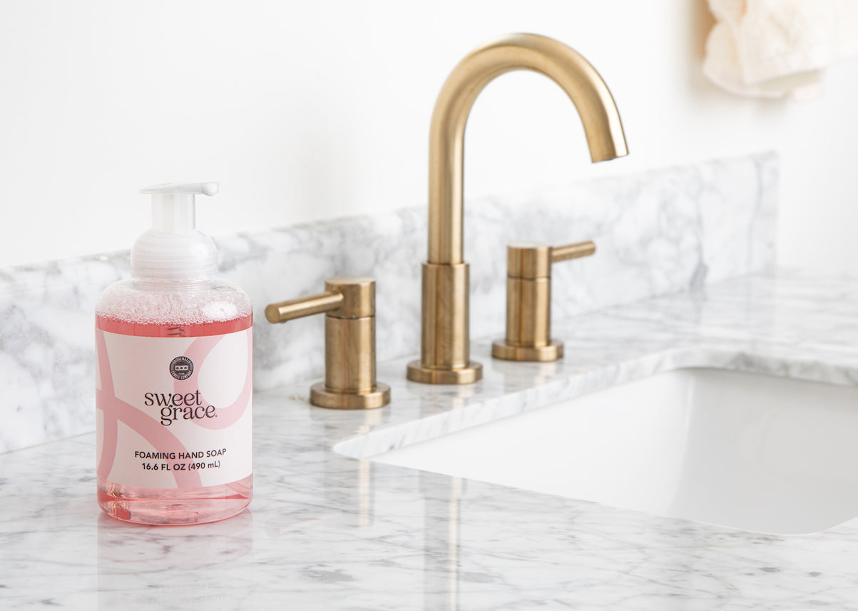 A bottle of Bridgewater Candle Company’s Sweet Grace Foaming Hand Soap, enriched with oriental fragrance and aloe, rests on a white marble bathroom countertop beside a brass faucet and handles.