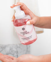 A person pumps Bridgewater Candle Company’s Sweet Grace Foaming Hand Soap from a clear bottle into their palm, with the pink liquid and label visible and a towel in the background, suggesting its signature oriental scent.