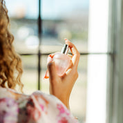 A woman with curly hair holding Sweet Grace spritz sweet grace perfume in the air with a window in the background