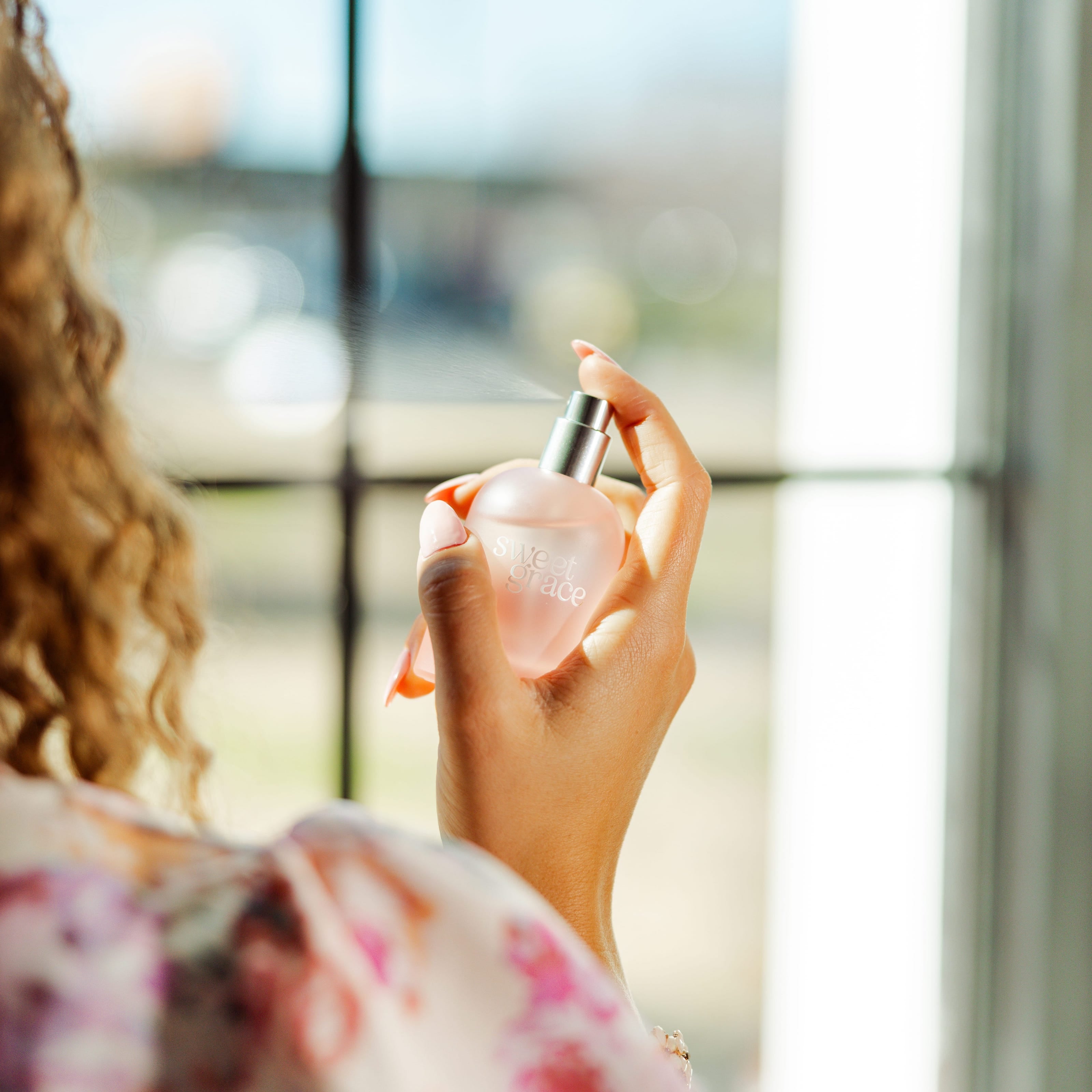 A woman with curly hair holding Sweet Grace spritz sweet grace perfume in the air with a window in the background