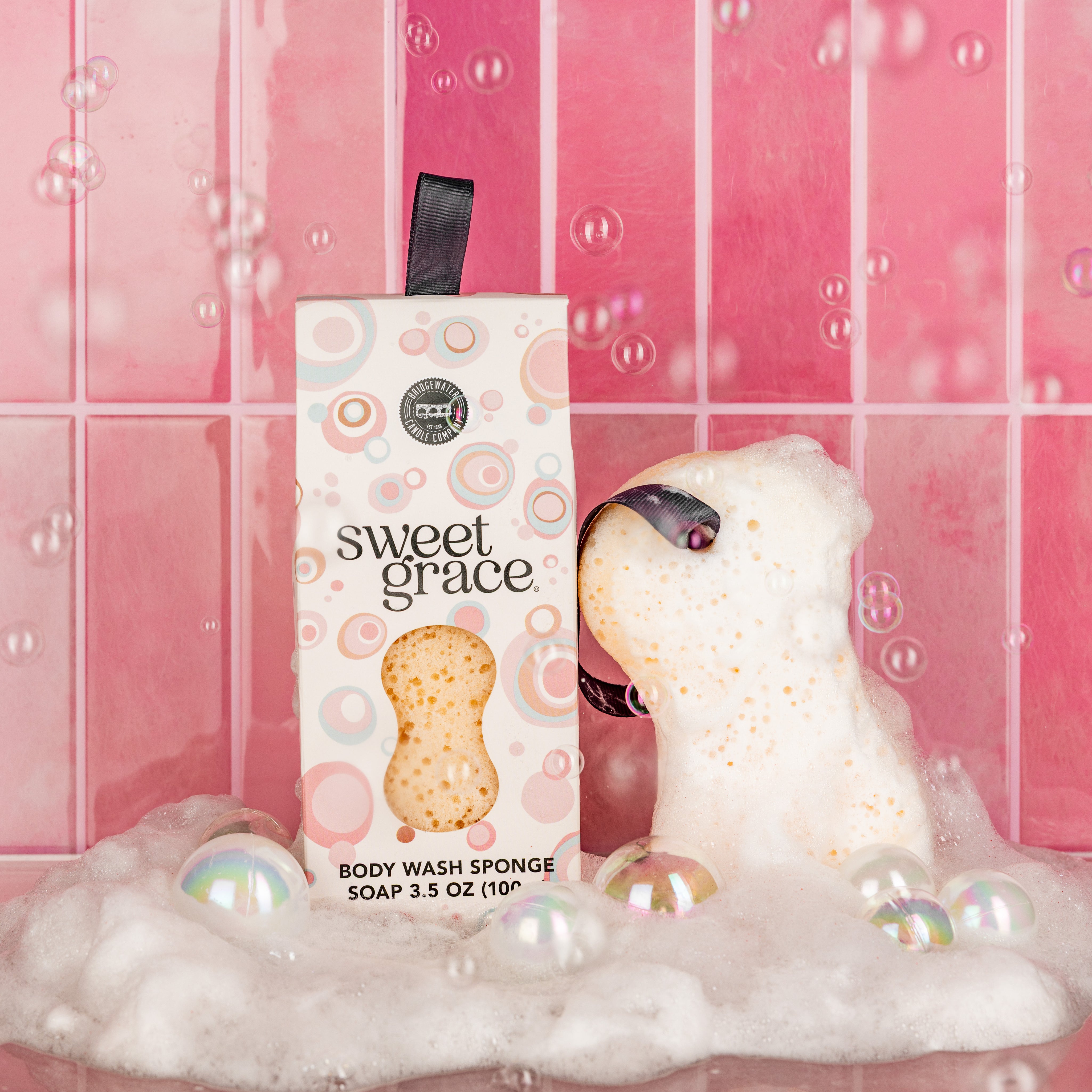 The Bridgewater Candle Sweet Grace Body Wash Sponge sits boxed on a soapy surface beside a bubbly dog-shaped sponge, ready to exfoliate, with floating bubbles and pink tiles in the background.