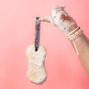 A hand with bracelets holds up the Bridgewater Candle Sweet Grace Body Wash Sponge by its strap against a pink background, ready to exfoliate and create a creamy lather.