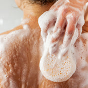 A close-up of a person washing their back with a soapy sponge, creating thick foam and bubbles on their skin. The scene focuses on the back and hand, with no face visible.