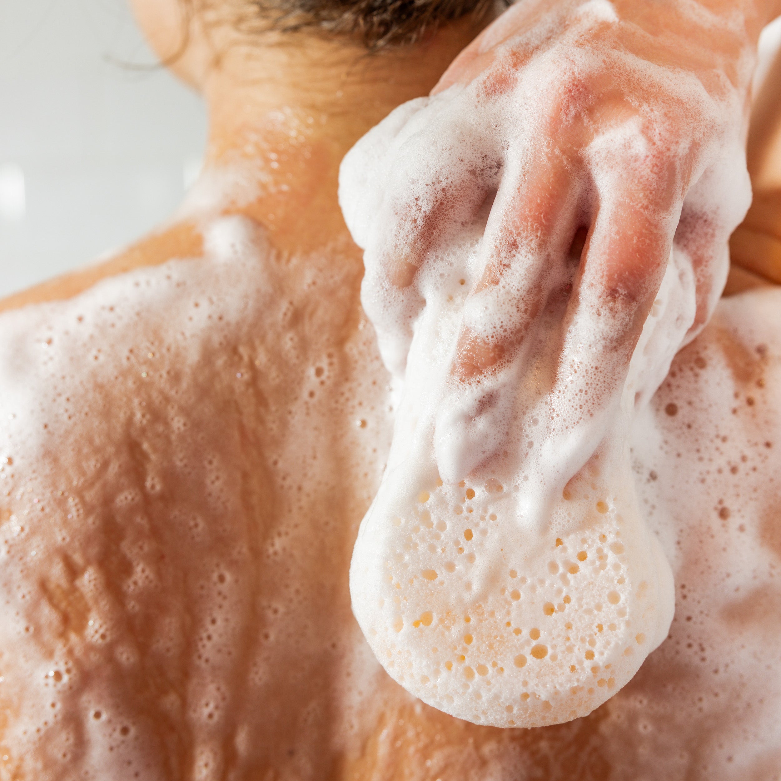 A close-up of a person washing their back with a soapy sponge, creating thick foam and bubbles on their skin. The scene focuses on the back and hand, with no face visible.