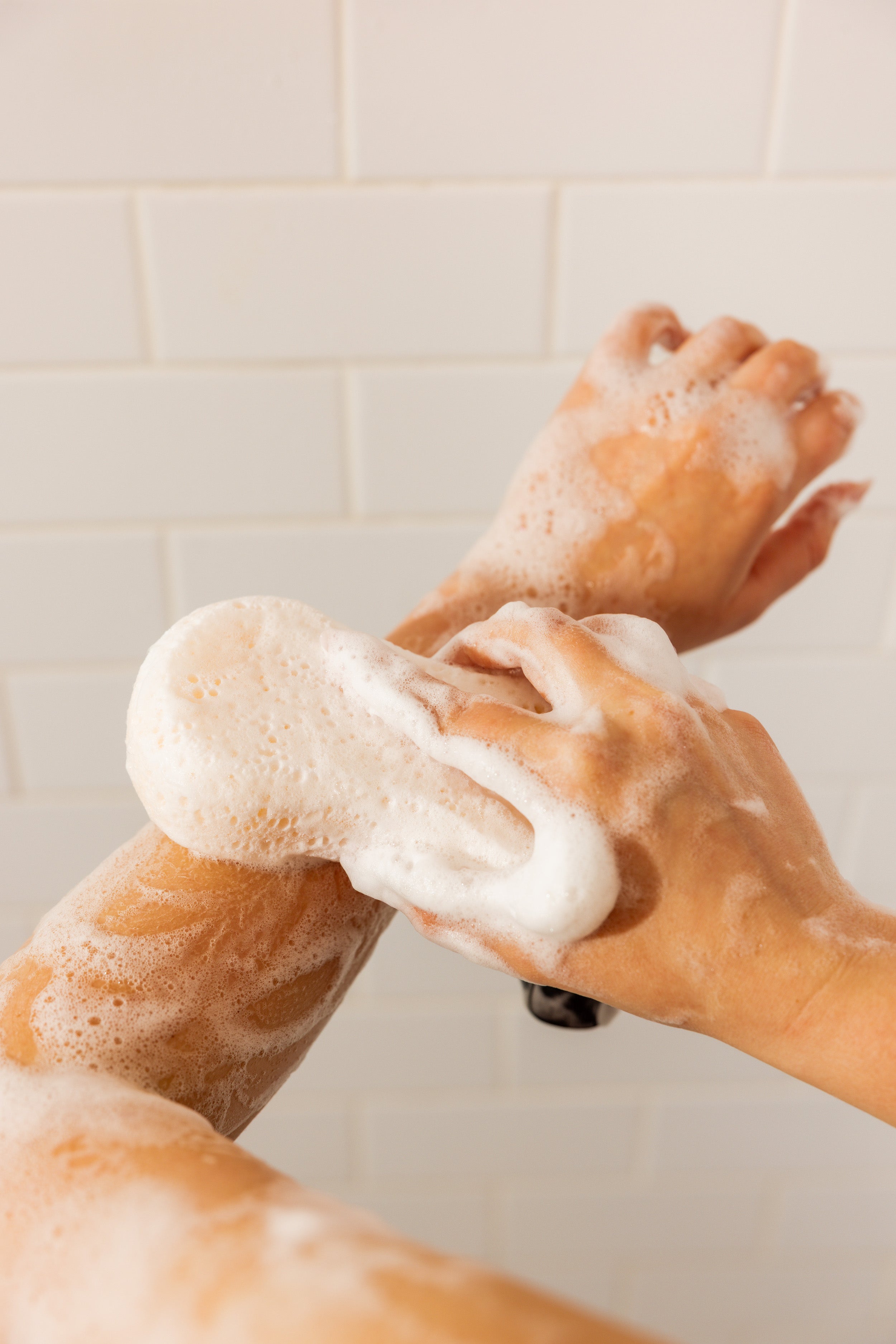 A person washing their arm with a white loofah filled with soap suds, against a background of white tiled wall.