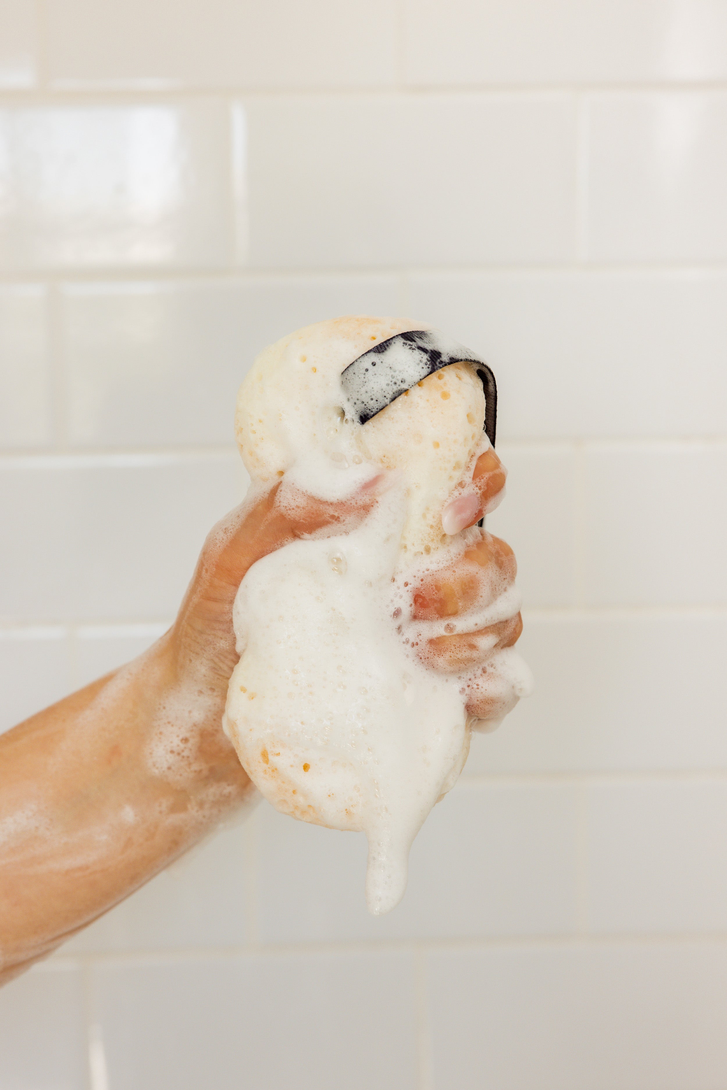 A hand squeezes a soapy, yellow bath sponge with white foam against a background of white tiled wall.