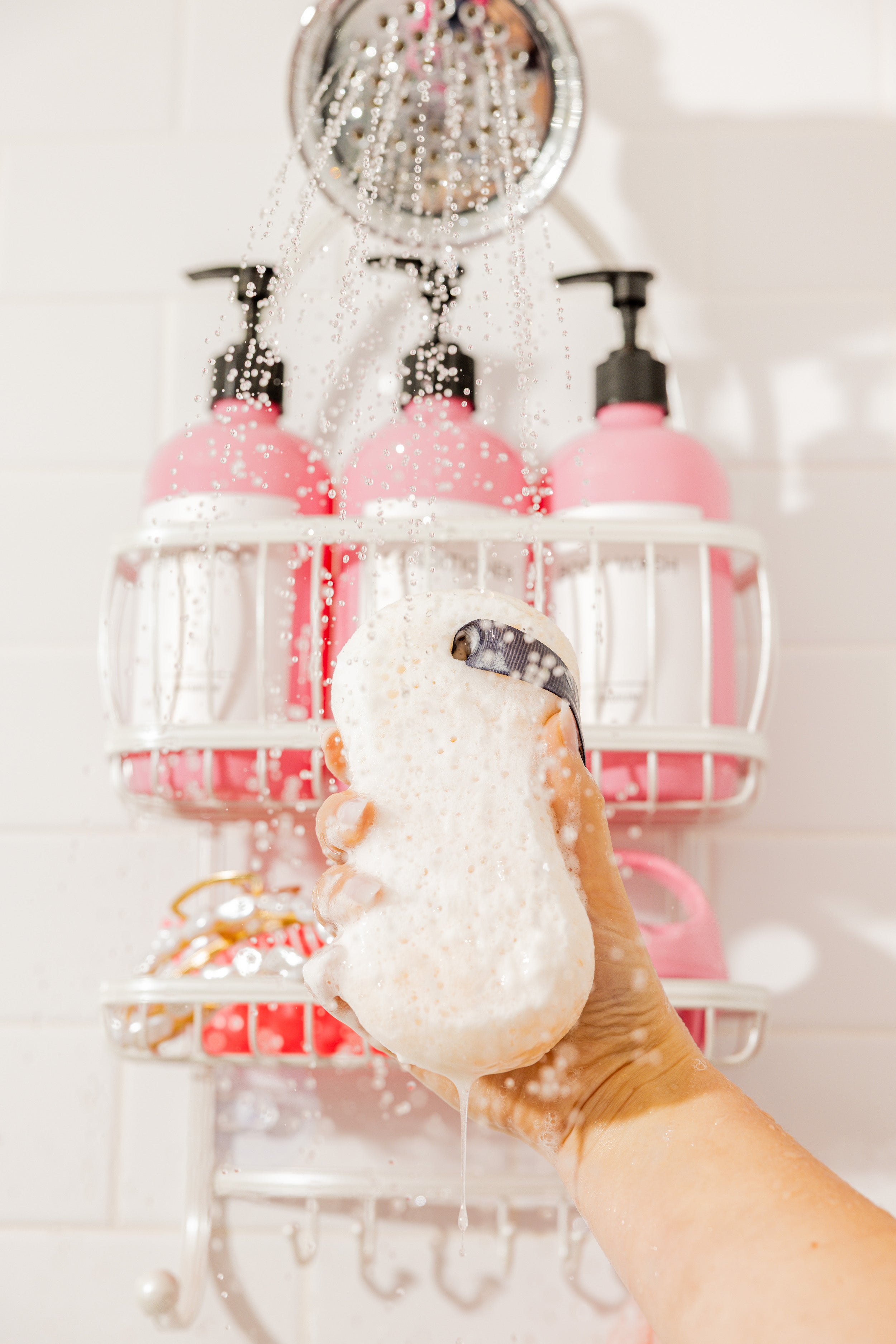 A hand holds a sudsy loofah sponge under a running showerhead, with pink bottles of bath products on a white shower caddy in the background. Water droplets are visible in the air.