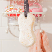 A close-up of the Sweet Grace Body Wash Sponge by Bridgewater Candle, hanging by a black ribbon with water droplets splashing—bathroom shelves and a peach towel in the background—perfect for exfoliating and creating a creamy lather.