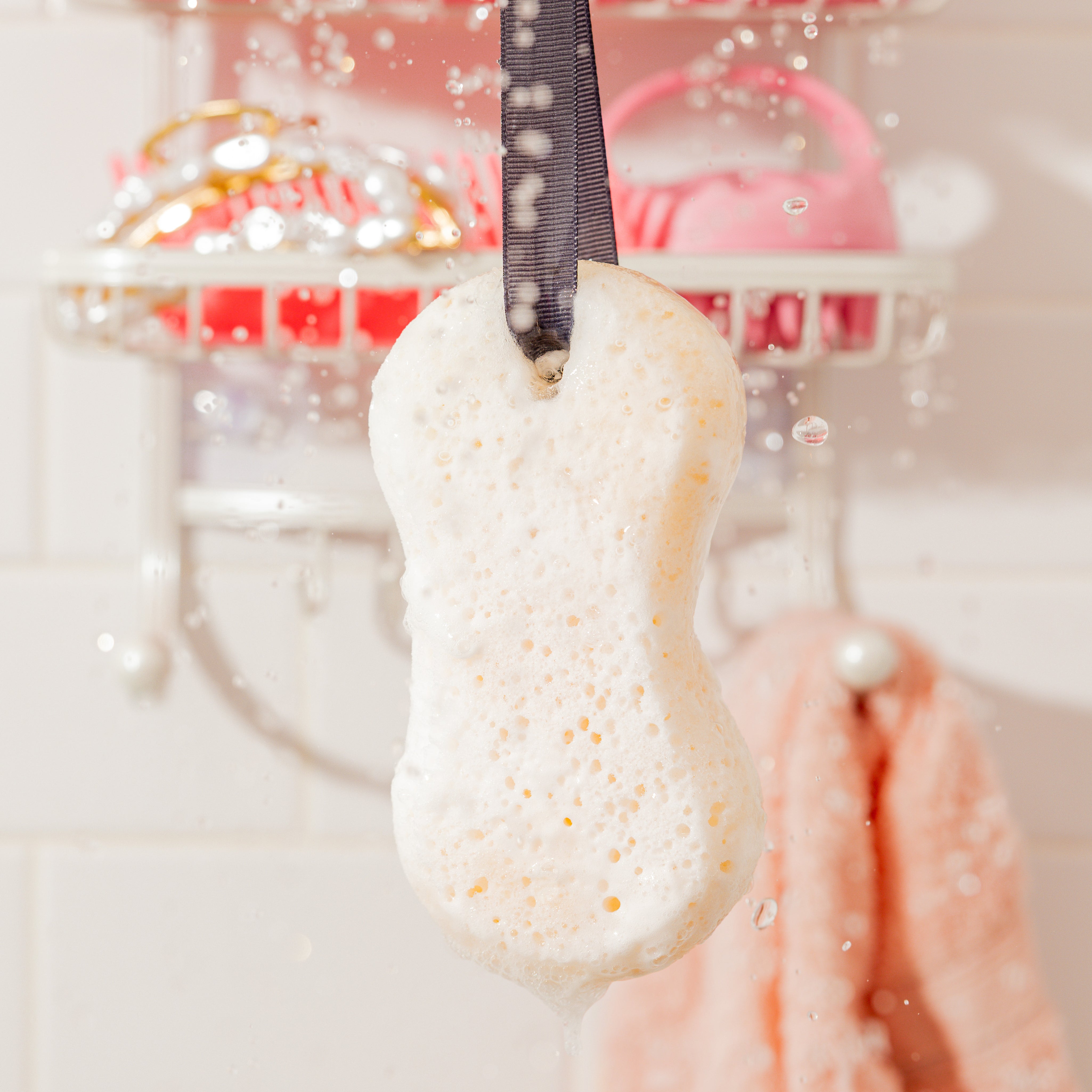 A close-up of the Sweet Grace Body Wash Sponge by Bridgewater Candle, hanging by a black ribbon with water droplets splashing—bathroom shelves and a peach towel in the background—perfect for exfoliating and creating a creamy lather.