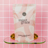 A pink and white bag of Bridgewater Candle Sweet Grace Soaking Salts with Pink Himalayan Sea Salt rests on a glass shelf with gold brackets against a pink tiled wall.