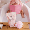 A woman relaxes in a bubble bath, wrapped in a pink towel and reading a pink journal. On a wooden tray are Bridgewater Candle Sweet Grace Soaking Salts, Pink Himalayan Sea Salt, and a pink bath pouf.
