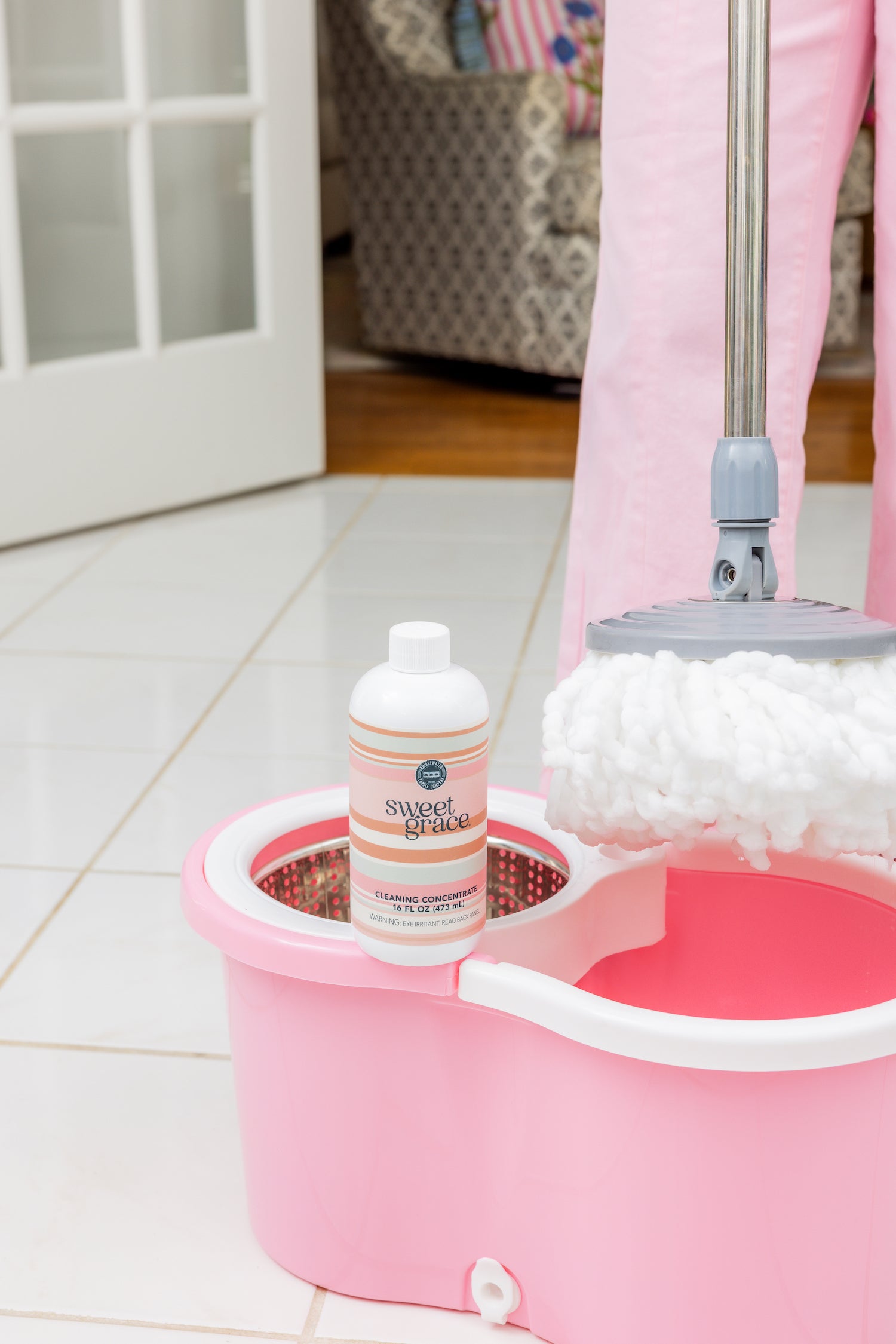 A pink mop bucket with a white mop and a bottle of Sweet Grace floor cleaner on top, set on a white tiled floor; a person in pink pants stands nearby.