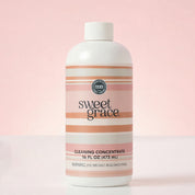 A bottle of Bridgewater Candle’s Sweet Grace Cleaning Concentrate | Mop Soap, a fragrant cleaner, stands against a soft pink background. The label displays the brand name, product name and type, size (16 fl oz), and a safety warning.